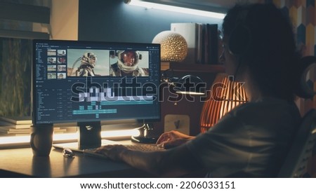 Male editor in headphones sitting at the table at night and editing video with astronauts for customer on pc in professional program while working remotely