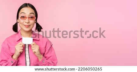 Portrait of stylish, modern asian girl, shows discount, credit card and look pleased, paying contactless, concept of shopping, standing over pink background