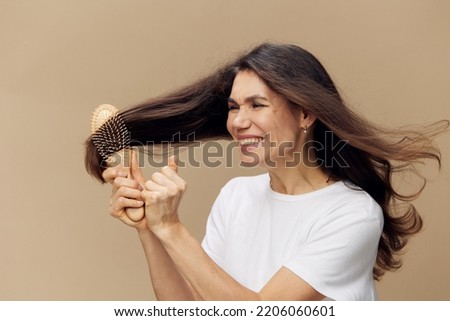 a sweet, beautiful middle-aged brunette woman stands pleasantly smiling on a beige background and combs her long hair with a wooden massage comb. Horizontal photo on