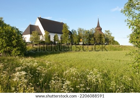 Frösö Church in evening lit. Flowers, meadows and farmland this side. Belfry to the right. Summer.