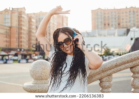 A young cheerful happy woman with dreadlocks, dressed in a white T-shirt, dancing, listening to music with headphones, resting, relaxing in a city park, walking along an alley. Urban lifestyle concept