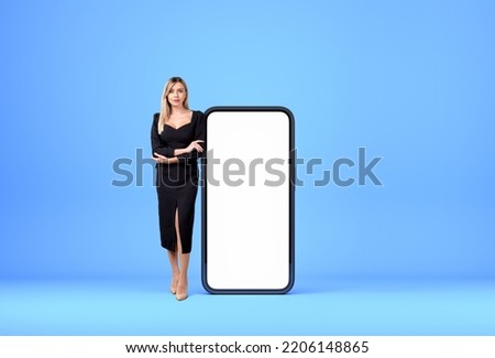 Businesswoman confident look, standing full length near large smartphone mock up empty screen, blue background. Concept of network and social media