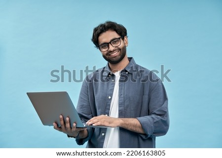 Happy young arab business man student or employee standing isolated on blue background holding laptop advertising web products for elearning, education training and webinars, working online.