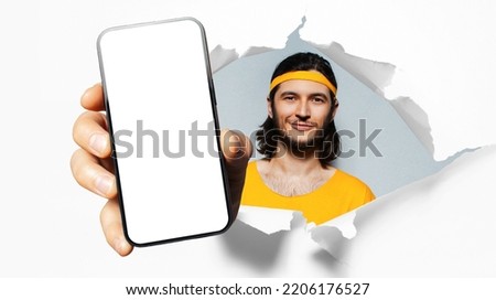 Studio portrait of young smiling sportsman, showing big smartphone with blank on screen through torn paper hole. White background. Wearing yellow band on head and shirt.