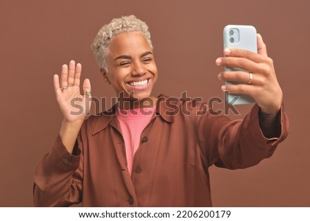 Young sociable smiling African American woman waving talking on video call on phone using messenger or popular application stands in studio with brown background. Mobile gadgets, smartphone