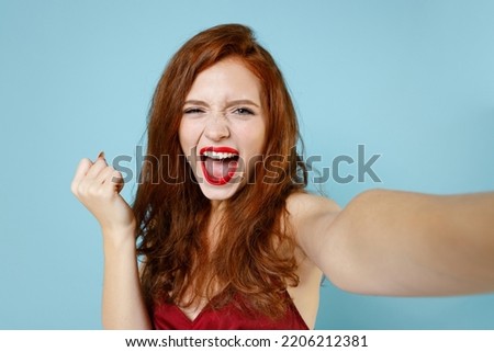 Close up young excited overjoyed redhead curly woman in red party dress doing selfie shot on mobile phone do winner gesture clench fist celebrating isolated on pastel blue background studio portrait