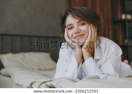 Young smiling fun woman wear white shirt pajama she lying in bed look camera prop up chin rest relax spend time in bedroom lounge home in own room hotel wake up dream be lost in reverie good mood day. Royalty-Free Stock Photo #2206212501