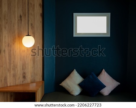 Empty white blank picture frame mockup hanging on blue wall background over the bench and pillows in the dark living room decorated with round ceiling lamp, full moon shape on wood wall.