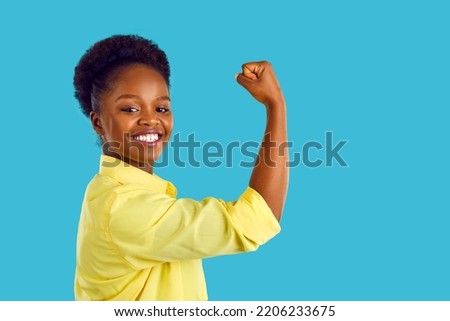 Side profile view of happy beautiful confident strong young Afro American woman in casual yellow shirt isolated on blue background smiling, flexing her arm and looking at camera. Girl power concept Royalty-Free Stock Photo #2206233675