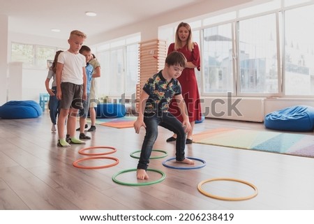 Small nursery school children with female teacher on floor indoors in classroom, doing exercise. Jumping over hula hoop circles track on the floor. Royalty-Free Stock Photo #2206238419