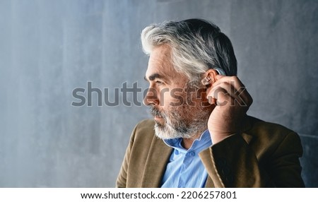 Grey-haired mature man with hearing impairment using hearing aid. Hearing solutions for deafness people Royalty-Free Stock Photo #2206257801