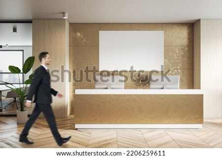 Businessman side view walking by modern reception area in golden color shades office with parquet floor and blank white poster on the wall with place for your logo or text, mock up