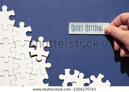 Quiet quitting text, caption. Jigsaw puzzle pieces half assembled and separate. Hand with sticky paper, office sticker. Top view, flat lay on monochromatic abstract blue white paper background. Royalty-Free Stock Photo #2206270761