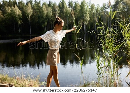 Picture of carefree girl hiker walking on toes along wild river with coniferous forest outstretching hands, enjoying fresh air, freedom, spending weekend outdoor far away from big noisy city