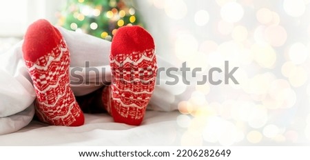 Cozy Merry Christmas morning at home. Kids feet in red warm socks covered with blanket on white bed near Christmas tree with bokeh lights. Happy moments of winter holiday. Background, copy space.