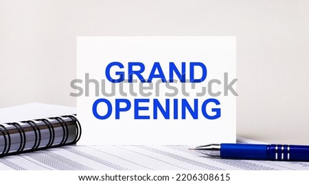 On a light gray background, a notebook, a blue pen and a sheet of paper with the text GRAND OPENING. Business concept