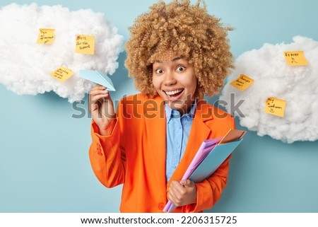 Positive businesswoman dressed in formal outfit holds folders and paper plane smiles gladfully enjoys starting on new working week writes down things to do on sticky notes isolated on blue background