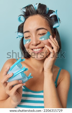 Vertical shot of pleased Asian woman makes hairstyle with hair rollers applies beauty patches under eyes undergoes skin care treatments dressed in striped t shirt isolated over blue background