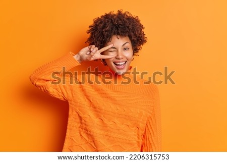 Indoor shot of happy curly haired woman makes peace gesture over eye shows v sign smiles toothily dressed in casual jumper isolated over vivid orange background. People body language and fun concept Royalty-Free Stock Photo #2206315753