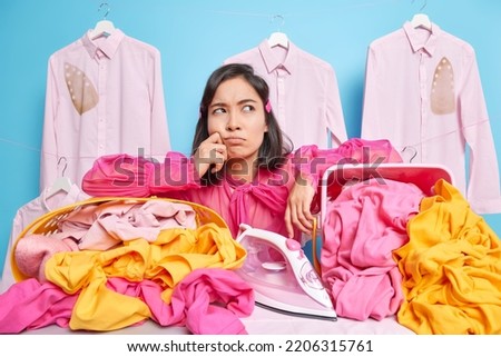 Thoughtful young Asian housewife brings huge pile of laundry on ironing board does household chores poses in washing room stands against blue background with hanging shirts. Cleaning day concept