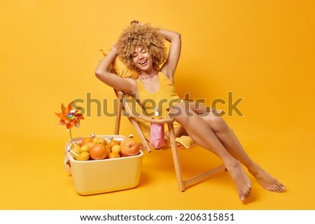 Positive curly haired young woman dressed in swim suit poses on comfortable deck chair laughs happily keeps eyes closed enjoys summer vacations at beach uses portabe fridge full of citrus fruits