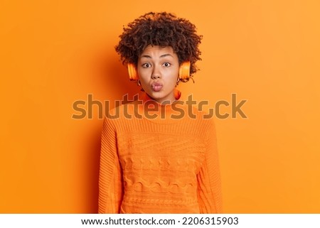 Horizontal shot of lovely curly haired woman keeps lips folded has romatic mood listens music via stereo headphones enjoys favorite song dressed in casual jumper isolated over orange background