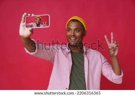 Gesturing V, victory or five handsome young african american man, guy making selfie using smartphone with back camera wearing pink shirt and yellow hat isolated on red background. 