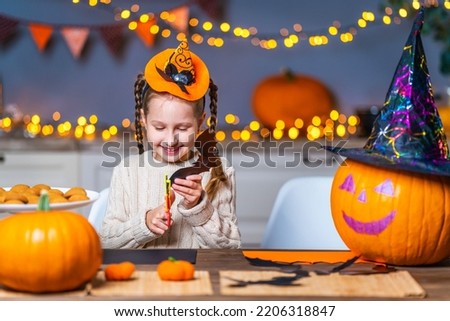 cute little girl with a hat on her head, sitting at a table in the kitchen at home, carves bats out of black cardboard, a symbol of the holiday. the child is smiling and getting ready for Halloween
