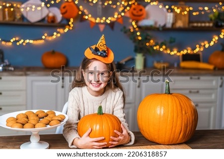 charming little girl with a hat on her head, sitting at kitchen table at home, holding and scaring a pumpkin for Halloween. symbol of the holiday. child is smiling and getting ready for Halloween