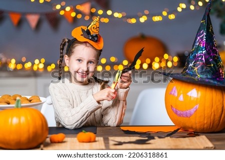 cute little girl with a hat on her head, sitting at a table in the kitchen at home, carves bats out of black cardboard, a symbol of the holiday. the child is smiling and getting ready for Halloween