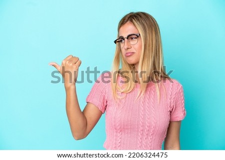 Blonde Uruguayan girl isolated on blue background unhappy and pointing to the side