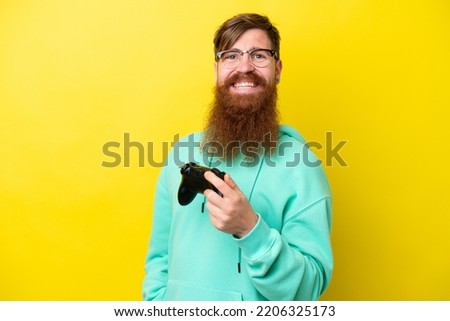 Redhead man with beard playing with a video game controller isolated on yellow background smiling a lot