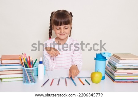 Indoor shot of smiling little schoolgirl with dark hair and braids sitting at table surrounded with books, having mathematics lesson, counting colored pencils, studying.