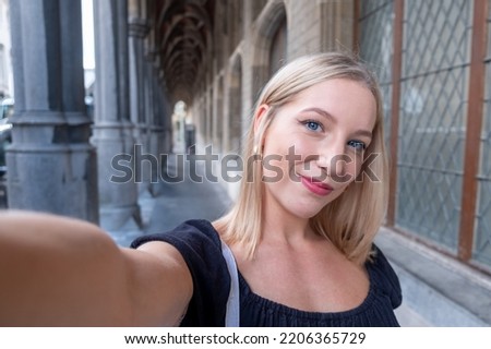 Portrait of wonderful white female model with bright makeup expressing energy in good day in Europe. Lovely blonde woman in stylish clothes making selfie while walking past old building. High quality