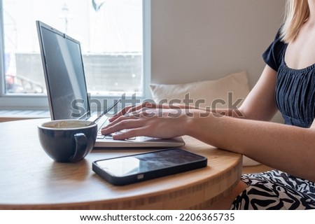 Close up side view of female using her laptop at a cafe. Lateral shot of young woman sitting at a table with a cup of coffee and mobile phone surfing the net on her laptop computer. High quality photo