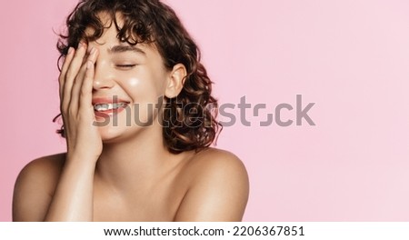 Haircare and beauty concept. Close up of happy smiling woman touches her curly natural hair, healthy glowing hair, white teeth and clean facial skin, standing against pink background.