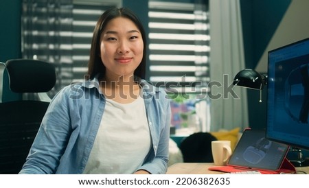 Female asian 3D designer sitting at the table near the modern computer and digital tablet, smiling and looking at camera while working on 3D project remotely from home.
