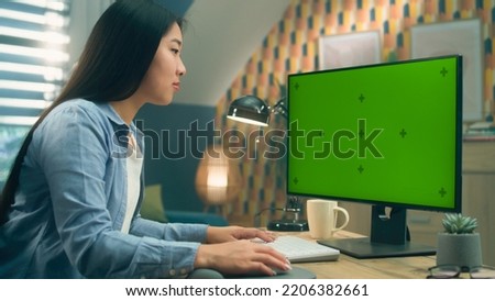 Asian woman sitting at the table and looking at screen of personal computer while working remotely from home or surfing the internet. Freelance. Green screen and chromakey