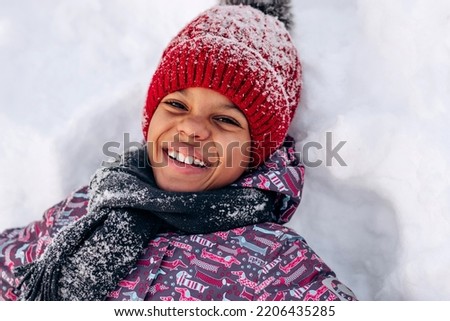 Happy little African-American girl in a red hat and jumpsuit is lying in the snow.Winter fun,active lifestyle concept.