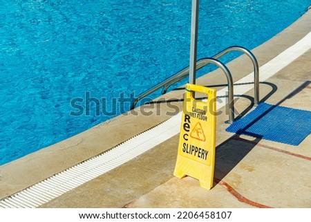 Safety sign with phrase Caution wet floor near the swimming pool at the tropical resort. Cleaning service slippery