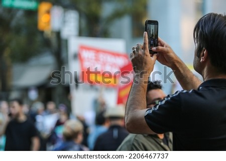 A man taking photos and videos with his phone at the blurry protest against in a daytime in a city 