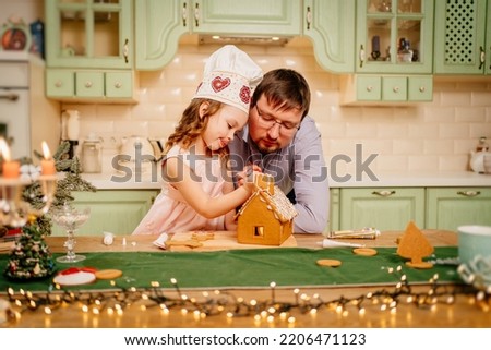 a cute little girl in a chef's cap with dad makes a gingerbread house with sugar glaze. traditional Christmas recipe. cooking school. happy family