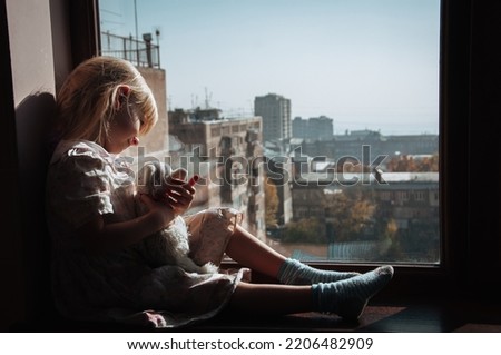 A girl with a toy is sitting on the windowsill. The child is bored.