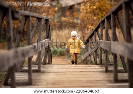 cute little child walks in nature on old park with wooden bridge on cold day. orange foliage, cold weather, warm trendy autumn bright tan and knitted clothes. autumn activities and family holidays