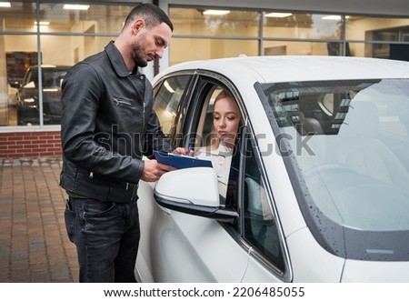 Young beautiful woman customer signing some important documents at new car dealership outdoors, sitting inside new white car. Handsome man holding agreement.