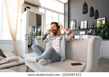 Positive bearded man listening music with headphones while sitting on couch at home