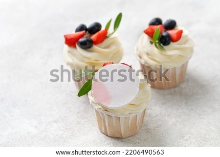 Cupcakes with fresh berries, vanilla cream, green leaves on grey background. Homemade dessert, mock up for picture. Round decor.