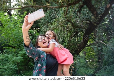 mother and daughter taking a self-portrait in the park, sitting on a bench and smiling