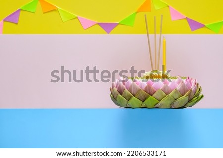 Selective focus at lotus pollen on top of pink lotus petal krathong with crown flower, incense stick and candle for Thailand Loy Krathong festival on colorful background with party flag.