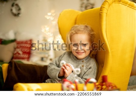 Cute little toddler curly girl in winter christmas sweater, sitting in cozy room, decorated for christmas